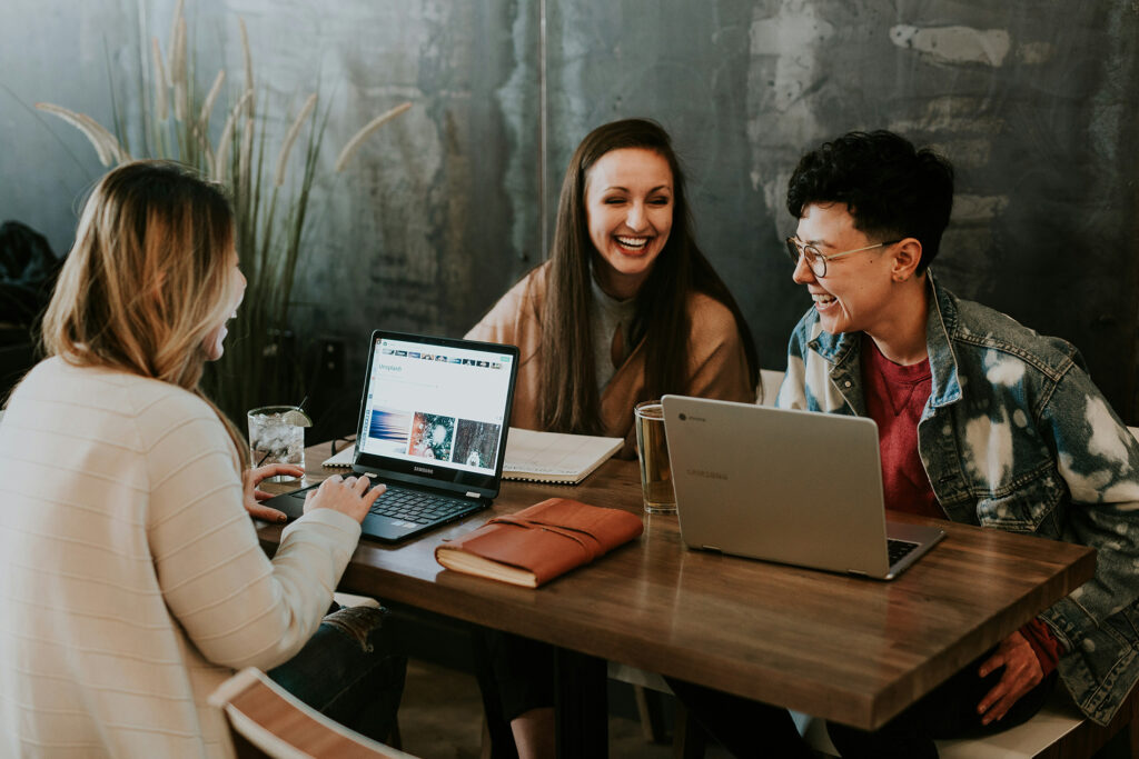 Três colegas de trabalho rindo juntas em volta de uma mesa com notebooks, exemplo de amizade no trabalho